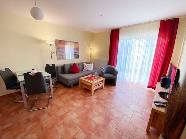 Uma sala de estar com sofá, poltronas, mesa de centro e mesa de jantar com cadeiras. Há um quadro na parede e um abajur.