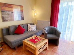 Uma sala de estar com um sofá cinzento, um travesseiro vermelho, uma mesa de centro de vidro e uma poltrona cinzenta.