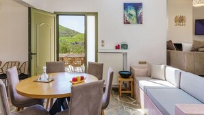 A dining area with a round table and chairs, a sofa, and a view of a green landscape through an open door. - Terra Divina Damnoni (Agios Vasileios)