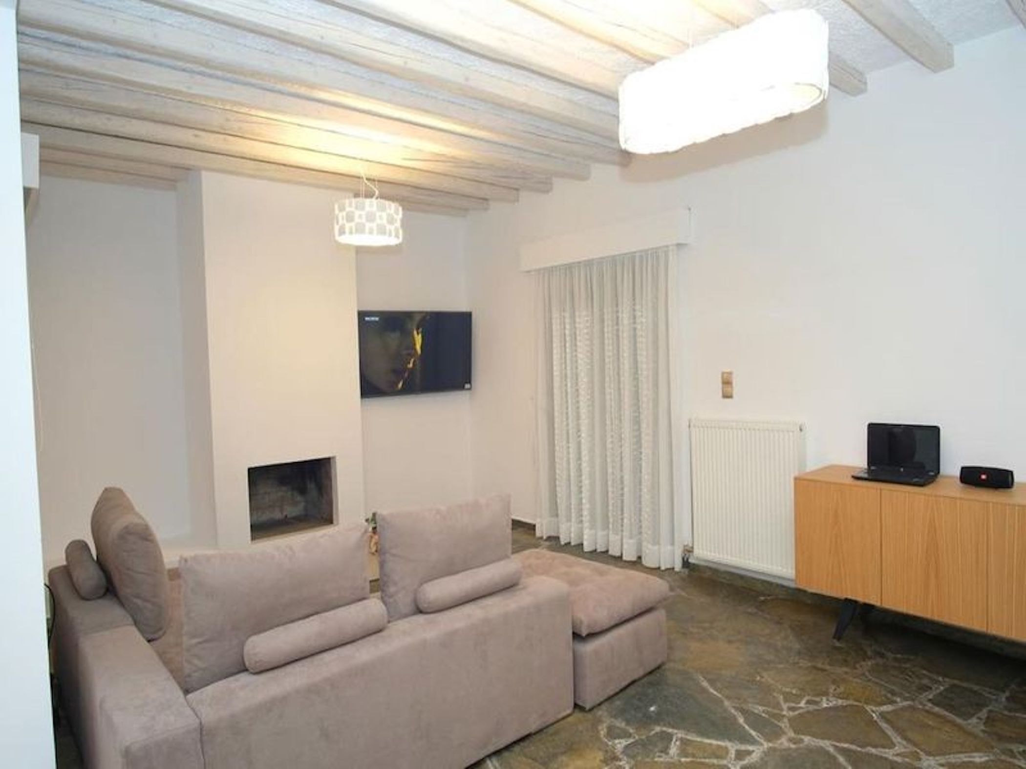 A living room with a beige sofa, a television mounted on the wall, a wooden cabinet, and a stone floor.