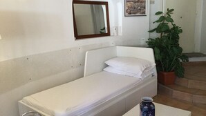 Ett enkelrum med en vit säng, ett litet bord, en vas, en krukväxt, en spegel som hänger på väggen och en inramad bild av en stadsbild.