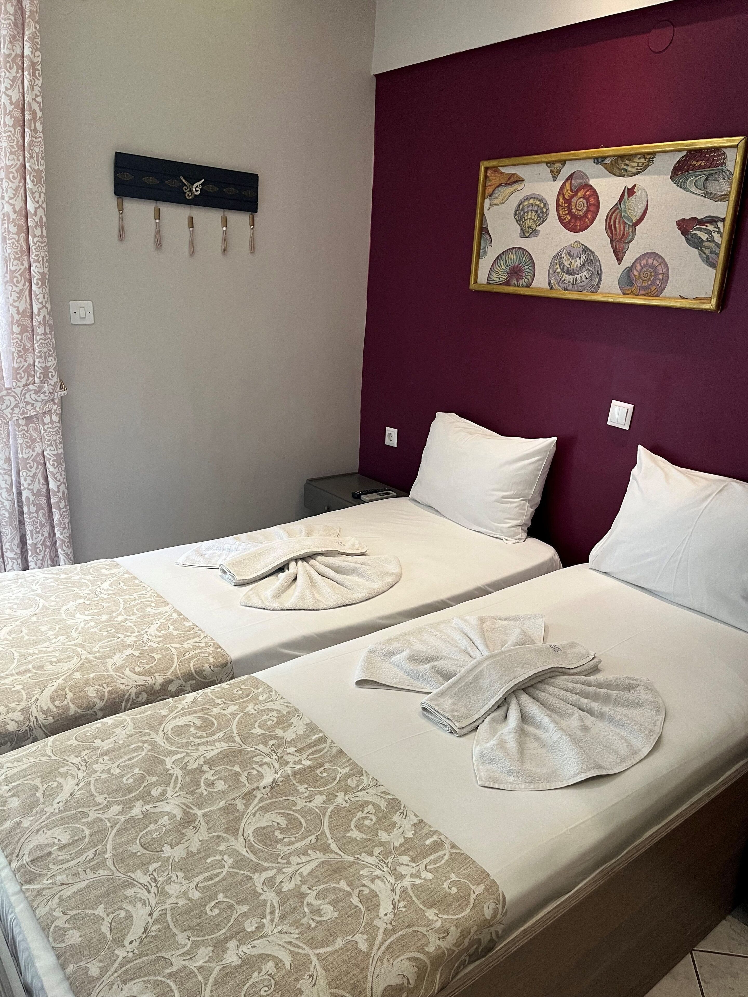 Hotellihuone, jossa on kaksi sänkyä. Jokaisessa sängyssä on kuviollinen peitto ja pyyhe. Seinälle on kiinnitetty hylly, jossa on koriste-esineitä, ja seinällä on myös kehystetty kuva simpukoista.