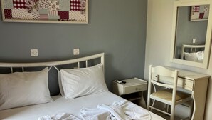 Una cama bien hecha con sábanas y toallas blancas, una mesita de noche con control remoto, un aparador con espejo y un colcha decorativa en la pared.