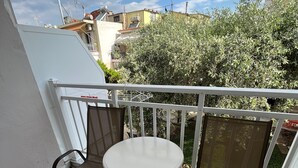 Un balcón con dos sillas y una mesa, barandilla blanca y vista a edificios y áreas verdes al fondo.