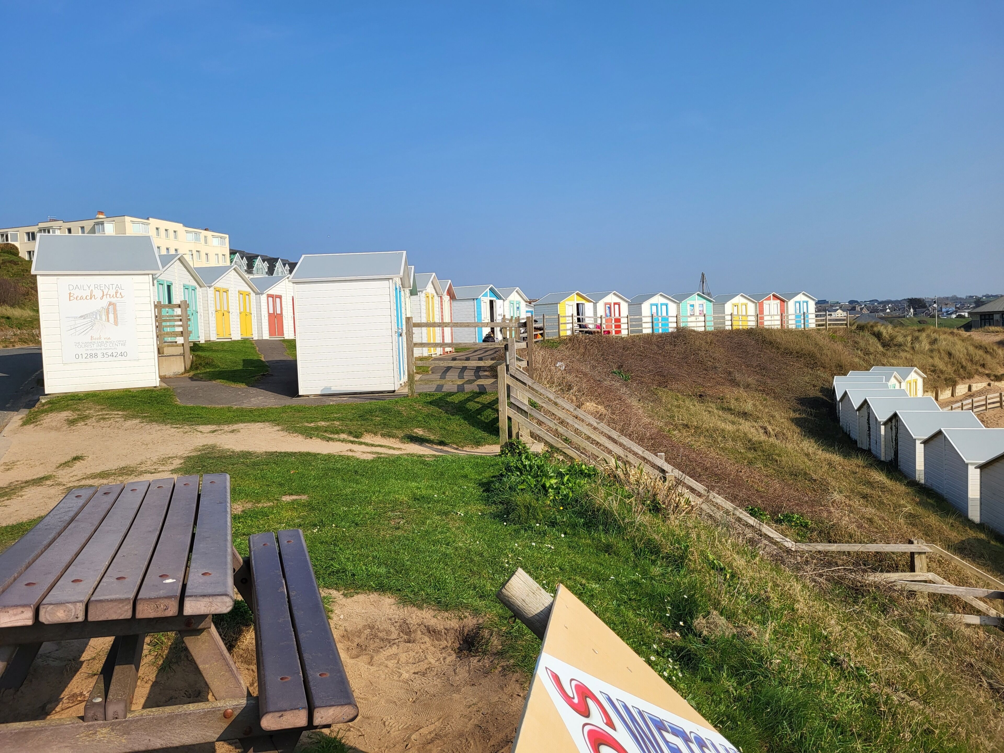 A row of beach huts with a wooden bench and a signboard in the foreground.