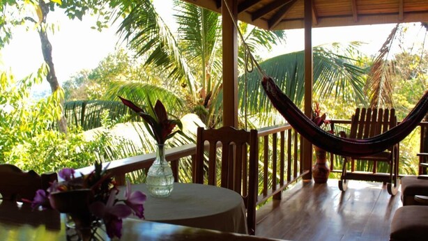 A wooden deck with a hammock, a table with a vase of flowers, and lush tropical foliage in the background. - Bungalow Casa Bosque, With Balcony and sea View (Drake Bay)