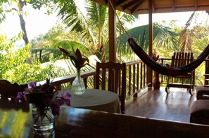 A wooden deck with a hammock, a table with a vase of flowers, and lush tropical foliage in the background. - Bungalow Casa Bosque, With Balcony and sea View (Drake Bay)