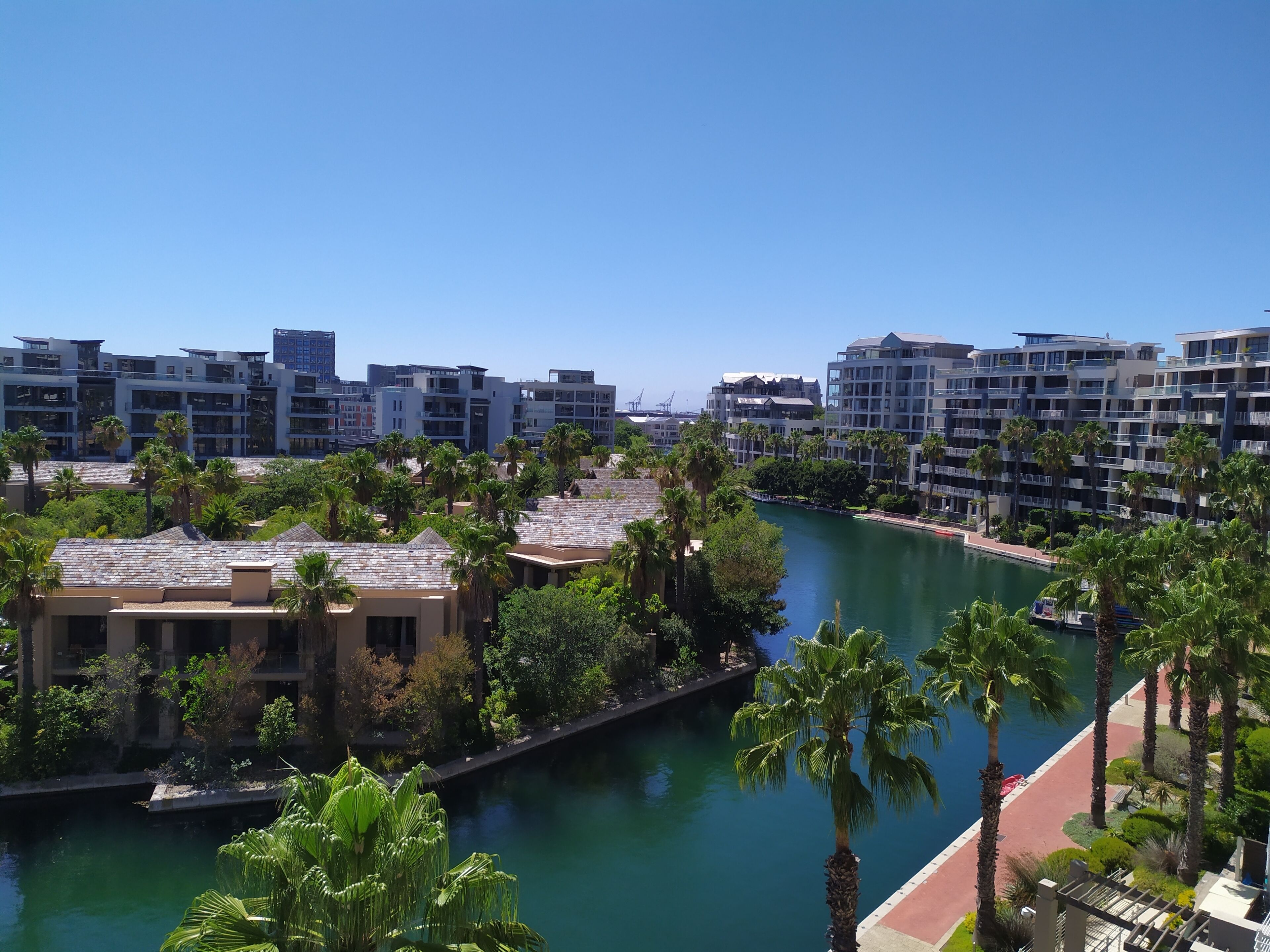 A riverside hotel with modern architecture, a water body, and palm trees.