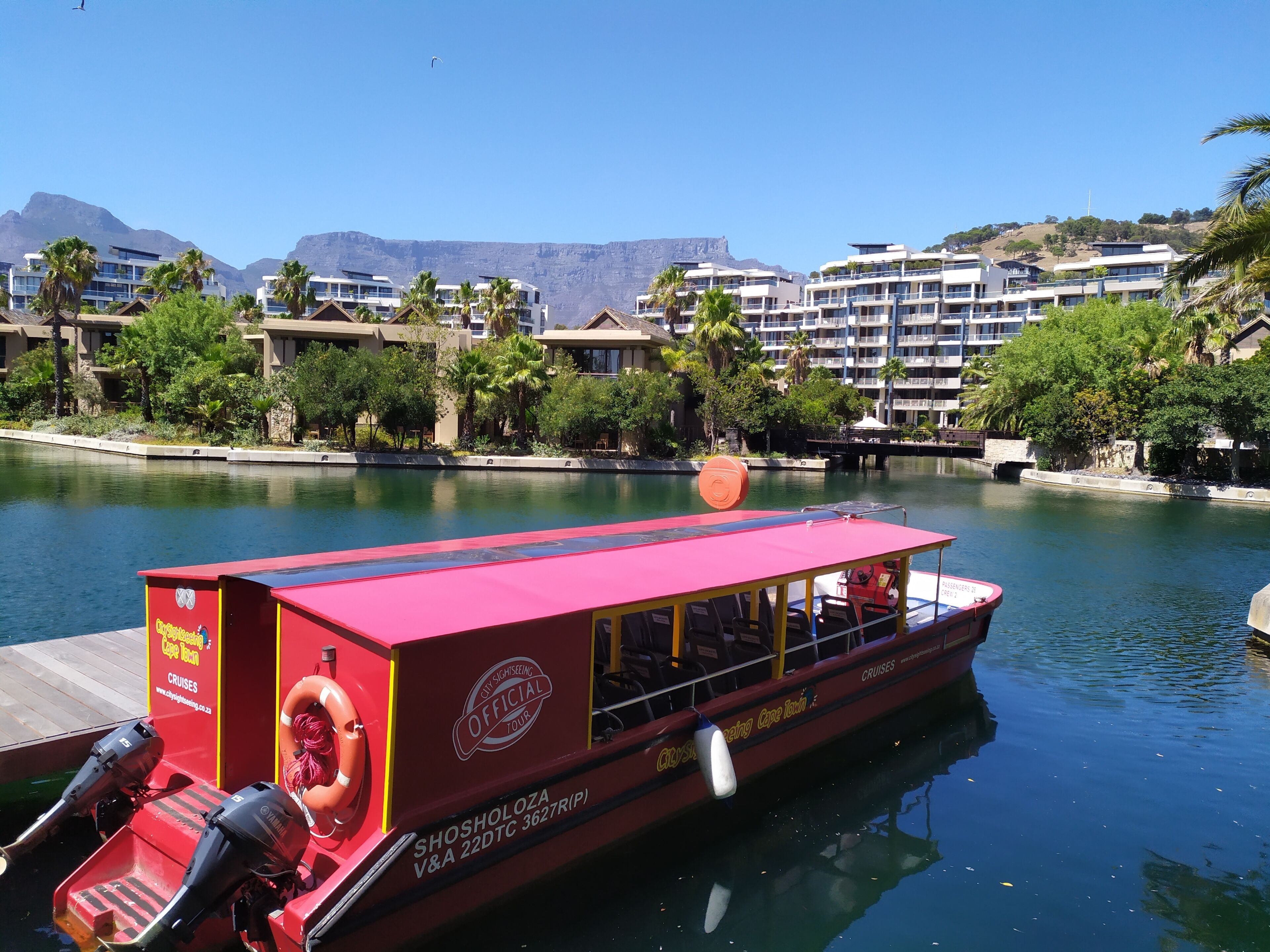 A red boat with "City Sightseeing Cape Town" written on it, docked at a marina with buildings and mountains in the background.