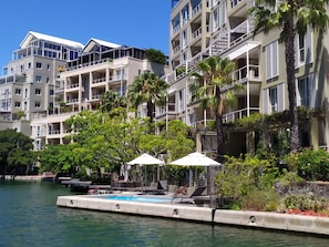 Un hotel frente al agua con varios balcones, una zona de piscina con tumbonas y abundante vegetación.