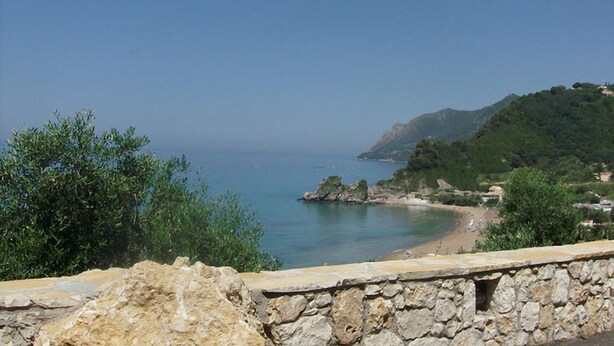 A coastal view with a stone wall in the foreground, a beach, and a hill with vegetation. - Holiday Studio Kontogialos in Pelekas Beach, Corfu (Corfu)