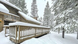 A snow-covered wooden deck with a railing, surrounded by snow-laden trees.