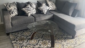 Ein modernes Wohnzimmer mit einem grauen Ecksofa, gemusterten Zierkissen, einem Glastisch und einem dekorativen Teppich.