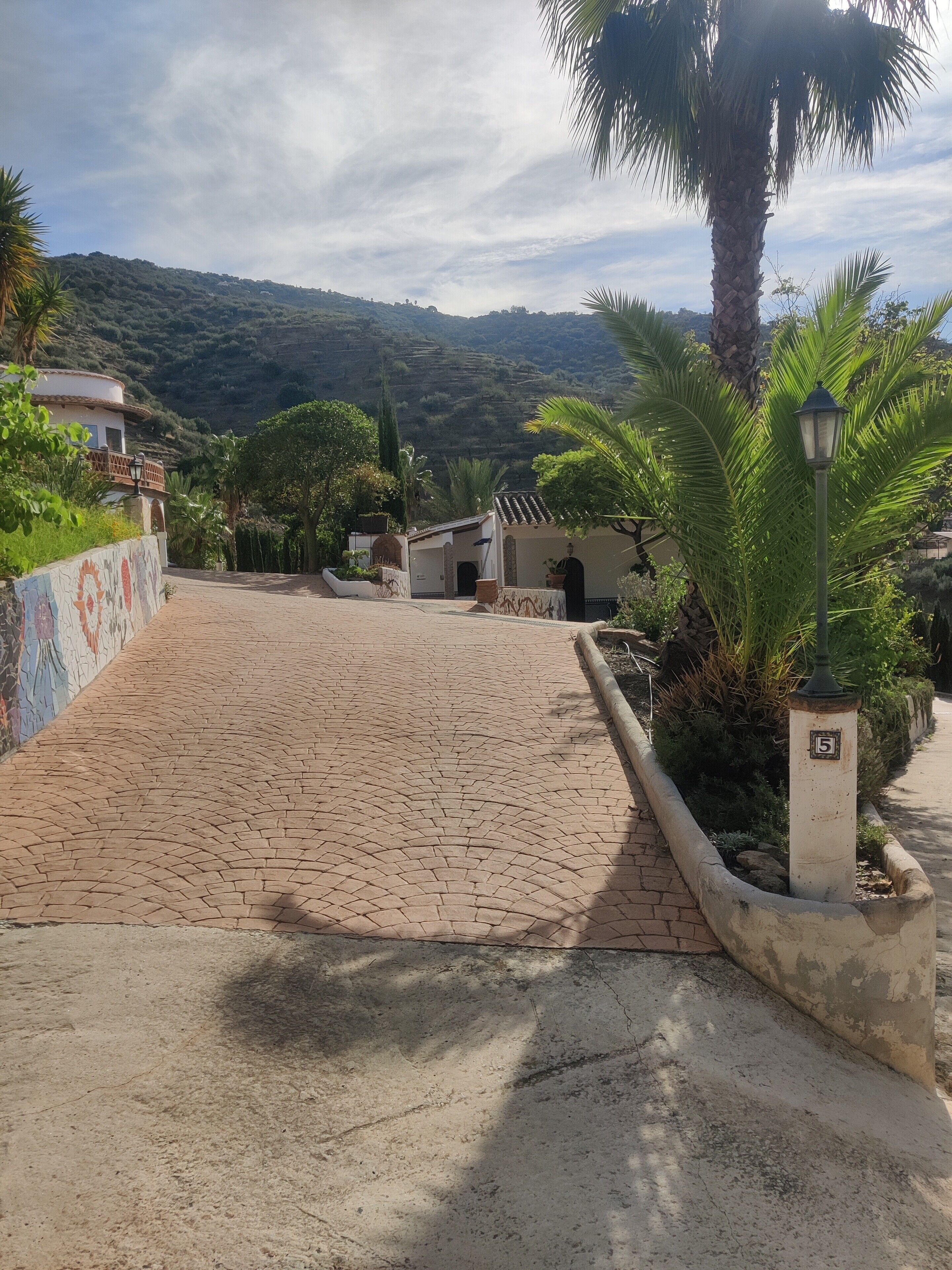 A paved pathway leading to a building with a tiled roof, surrounded by greenery and a mountainous backdrop.