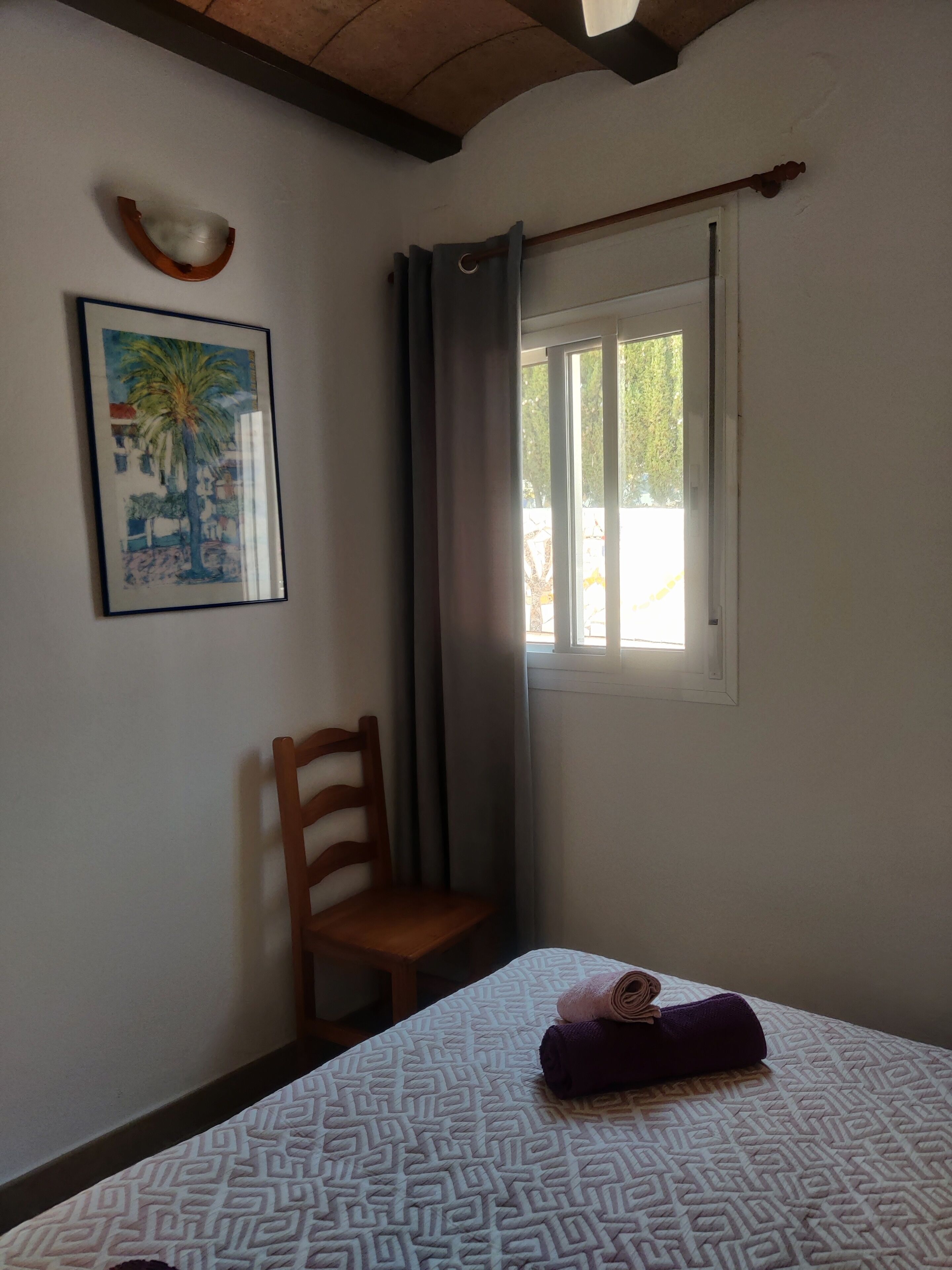 Een kamer met een bed, een stoel, een raam met gordijnen, een schilderij en een kleine tafel met een kom.