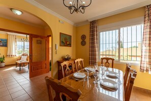 A dining room with a wooden table set for four, a chandelier, and a view through a window with curtains.