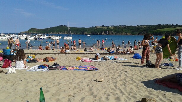 Una playa con gente tomando el sol y nadando, barcos en el agua y un paisaje montañoso al fondo.