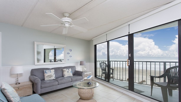 Ein geräumiger Balkon mit Blick auf den Strand, ein graues Sofa, ein Korbstuhl und ein Deckenventilator.