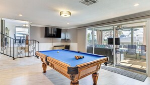 Una mesa de billar en una habitación bien iluminada, con un televisor montado en la pared, una escalera y vistas al exterior a través de amplias puertas correderas de vidrio.
