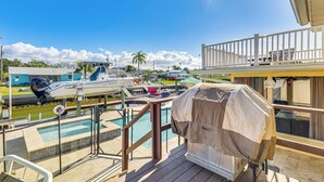 Zona de piscina con una parrilla cubierta, terraza de madera y vistas a barcos y edificios al fondo.