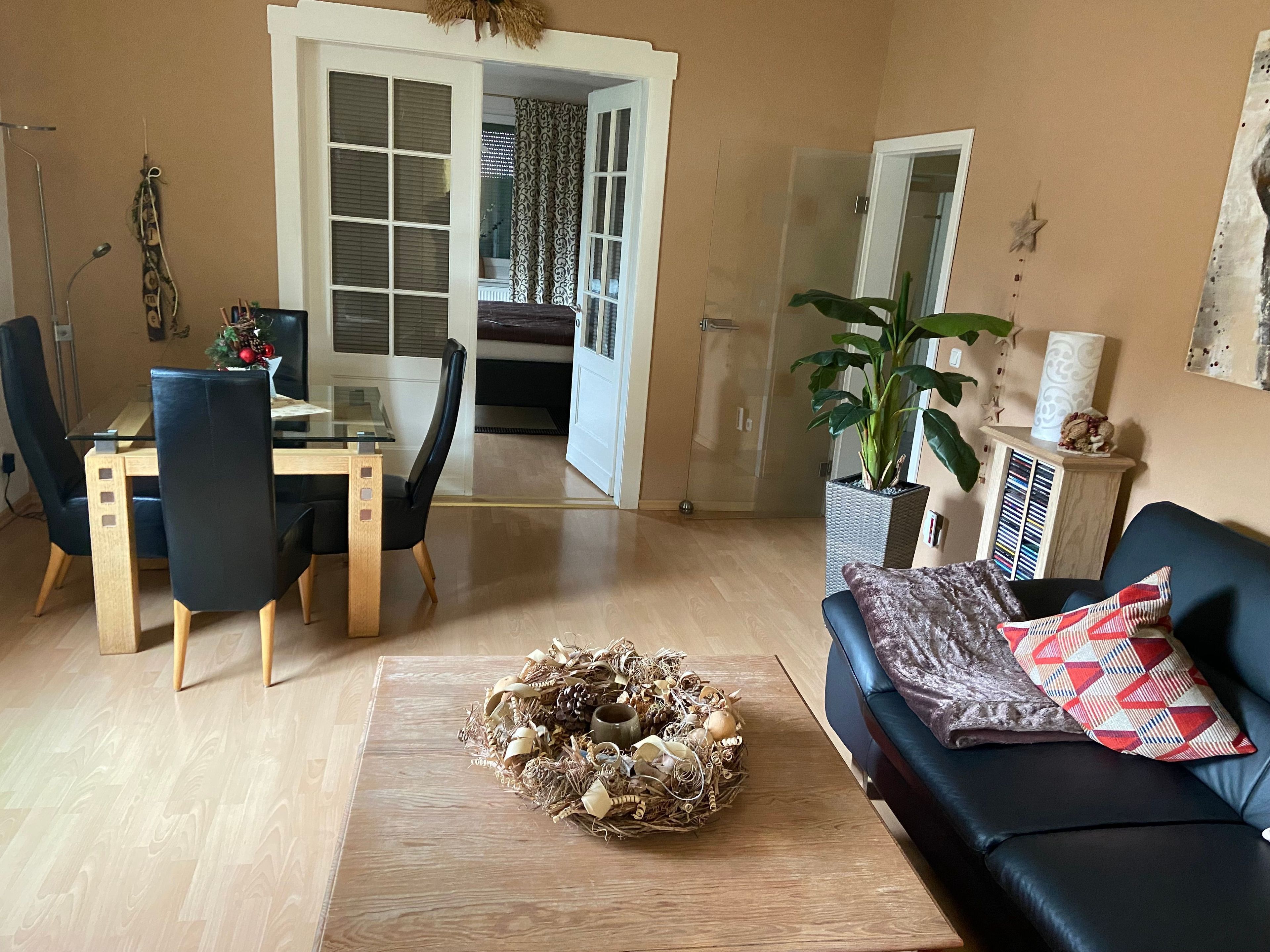 Ein Wohnzimmer mit einem Glastisch, schwarzen Stühlen, einer schwarzen Couch, einem dekorativen Kranz und einer großen Topfpflanze.