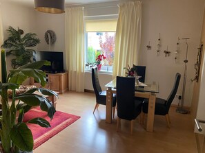Ein Wohnzimmer mit Fernseher, Esstisch, Stühlen, einem roten Teppich und einer großen Pflanze.