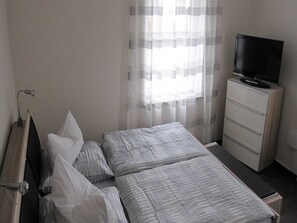 Ein Zimmer mit einem Bett, einem Fernseher auf einer Kommode und einem Fenster mit durchsichtigen Vorhängen.