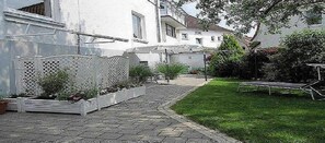 Ein gepflasterter Weg führt zu einem weißen Gebäude mit einem Gitterzaun und einem kleinen Garten.
