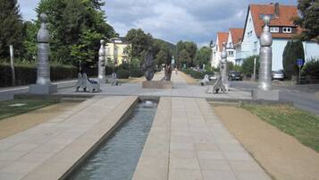 A paved pathway with a central water feature, flanked by sculptures and trees, leading to residential buildings.