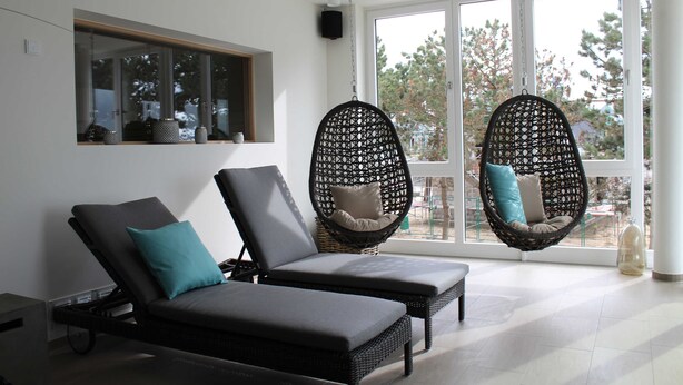 A modern room with two black lounge chairs, one with a teal pillow, and two hanging chairs with cushions, all facing a large window with a view of trees. - Apartment Seaside - Whg. D-05, 3 Zi., 2 Bäder, Südbalkon (Neustadt in Holstein)