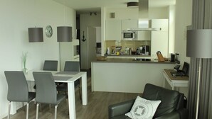 A modern kitchen with white cabinets, a microwave, and a coffee maker. There is a dining area with a white table and chairs, and a living area with a black leather sofa. - Apartment Seaside - Whg. D-05, 3 Zi., 2 Bäder, Südbalkon (Neustadt in Holstein)