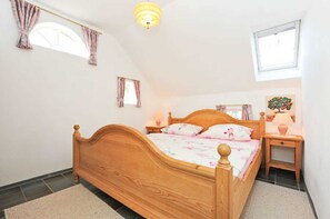 Un dormitorio con cama de madera, una mesita de noche con lámpara, una pequeña mesa con un jarrón y una ventana con cortinas.
