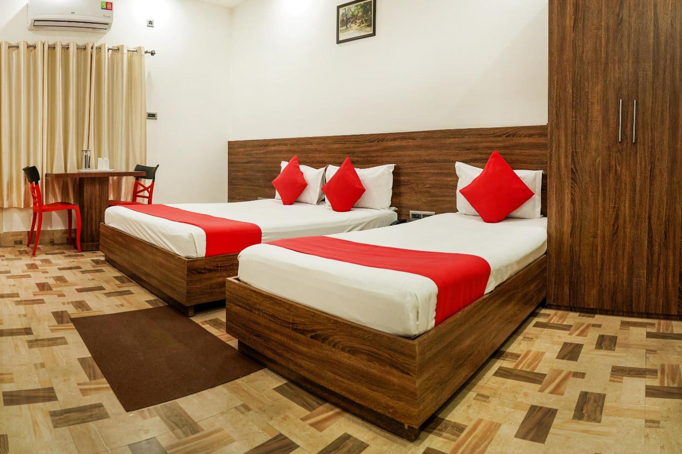 A hotel room with two wooden beds, each with red bedding and pillows, a small wooden table with chairs, and a brown rug on the floor.