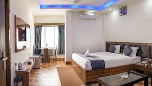 A hotel room with a bed, bedside table, desk, chair, and a view of the outside. - Hotel Heritage (Cooch Behar)