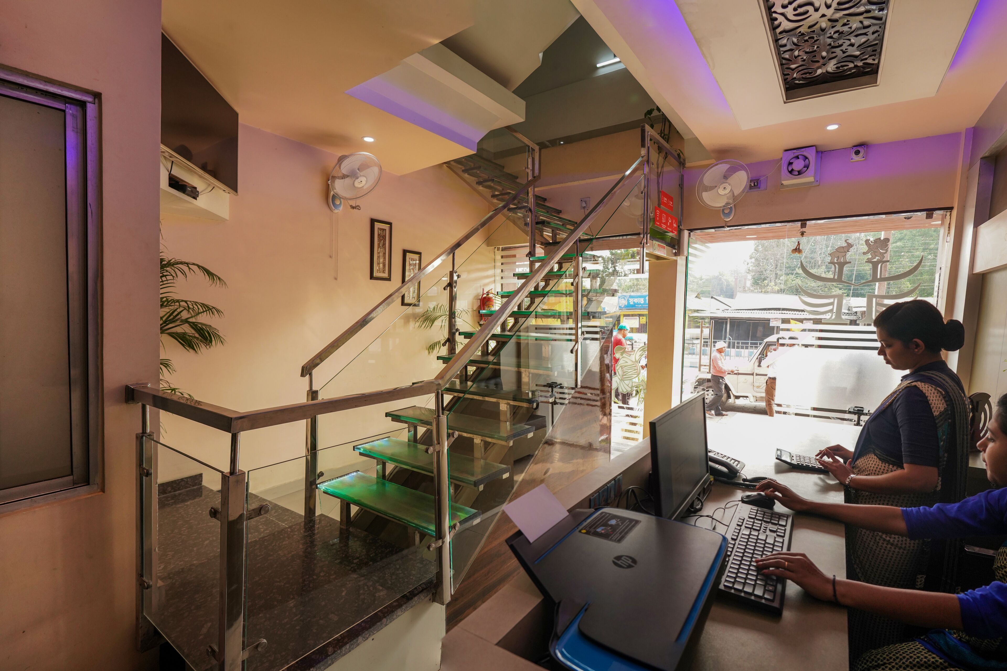 A modern interior with a staircase, glass railings, and a desk area with two people working on laptops.