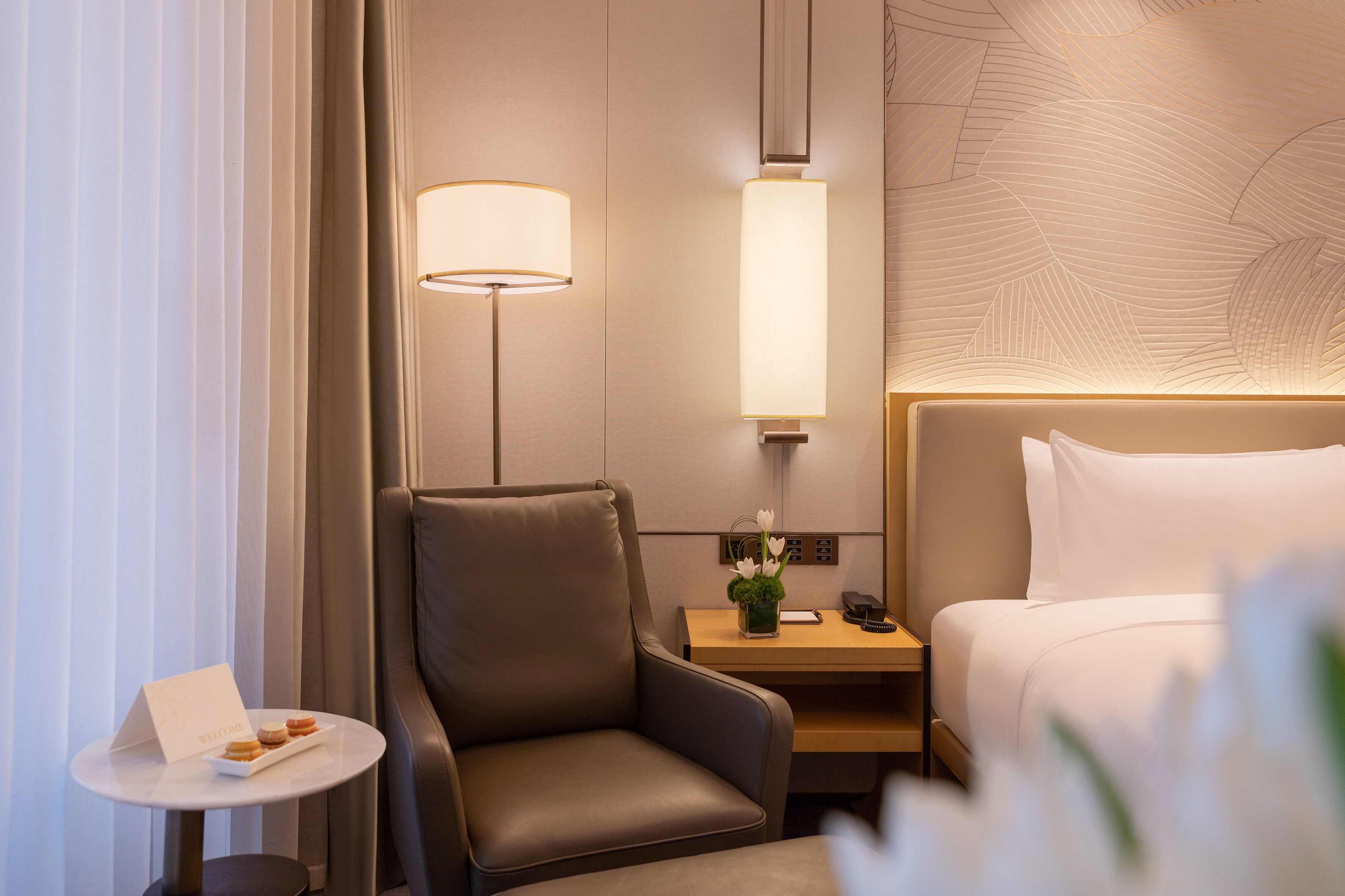 A hotel room with a bed, a desk, a chair, a nightstand, a lamp, and a small table with a card and a flower.