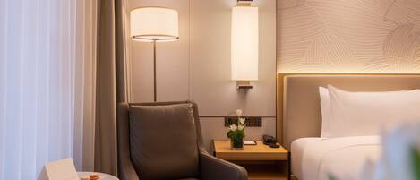 A hotel room with a bed, a desk, a chair, a nightstand, a lamp, and a small table with a card and a flower.