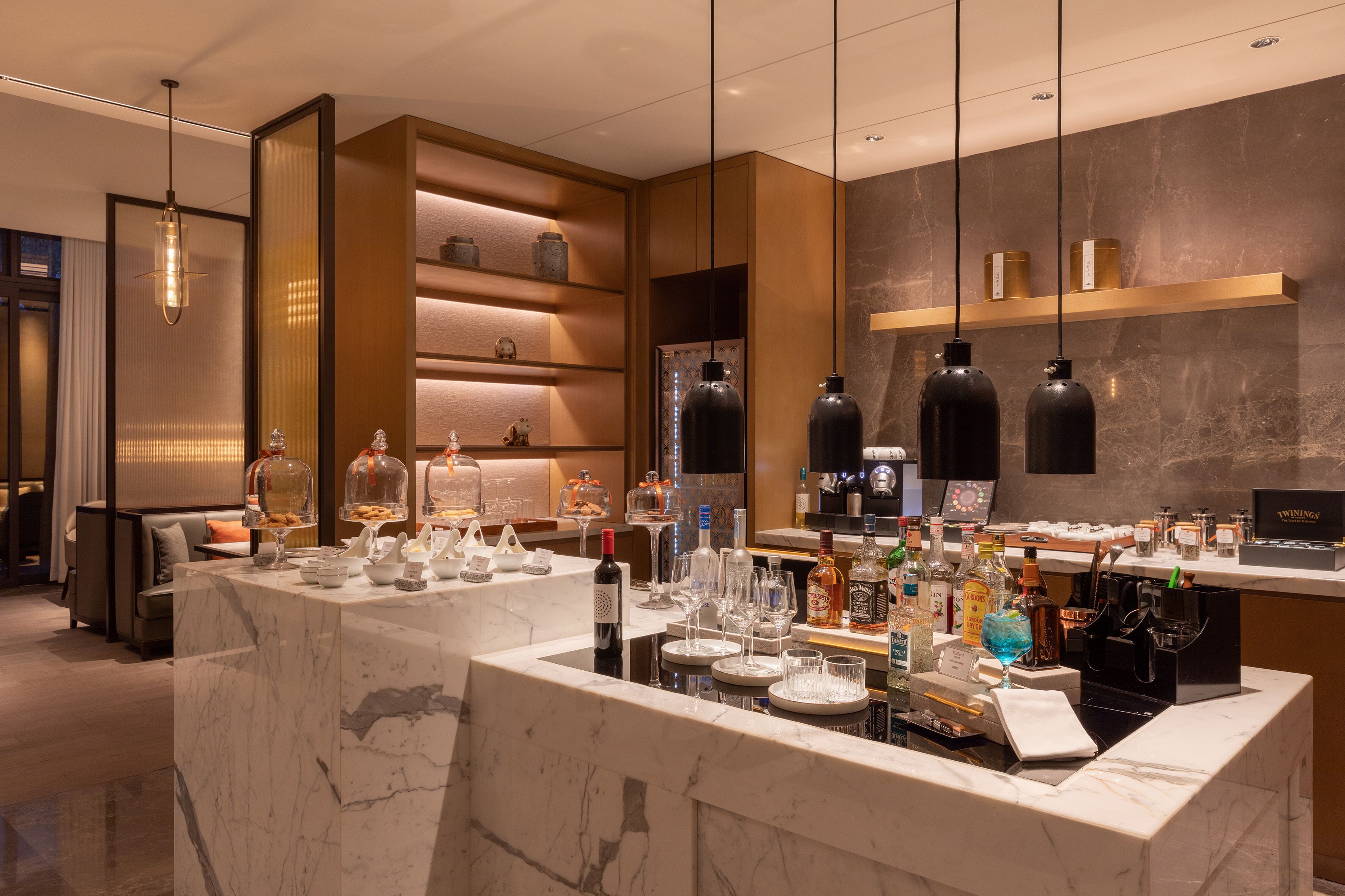 A modern bar area with marble countertops, wine glasses, and bottles. There are shelves with decorative items and a seating area with a sofa and cushions.