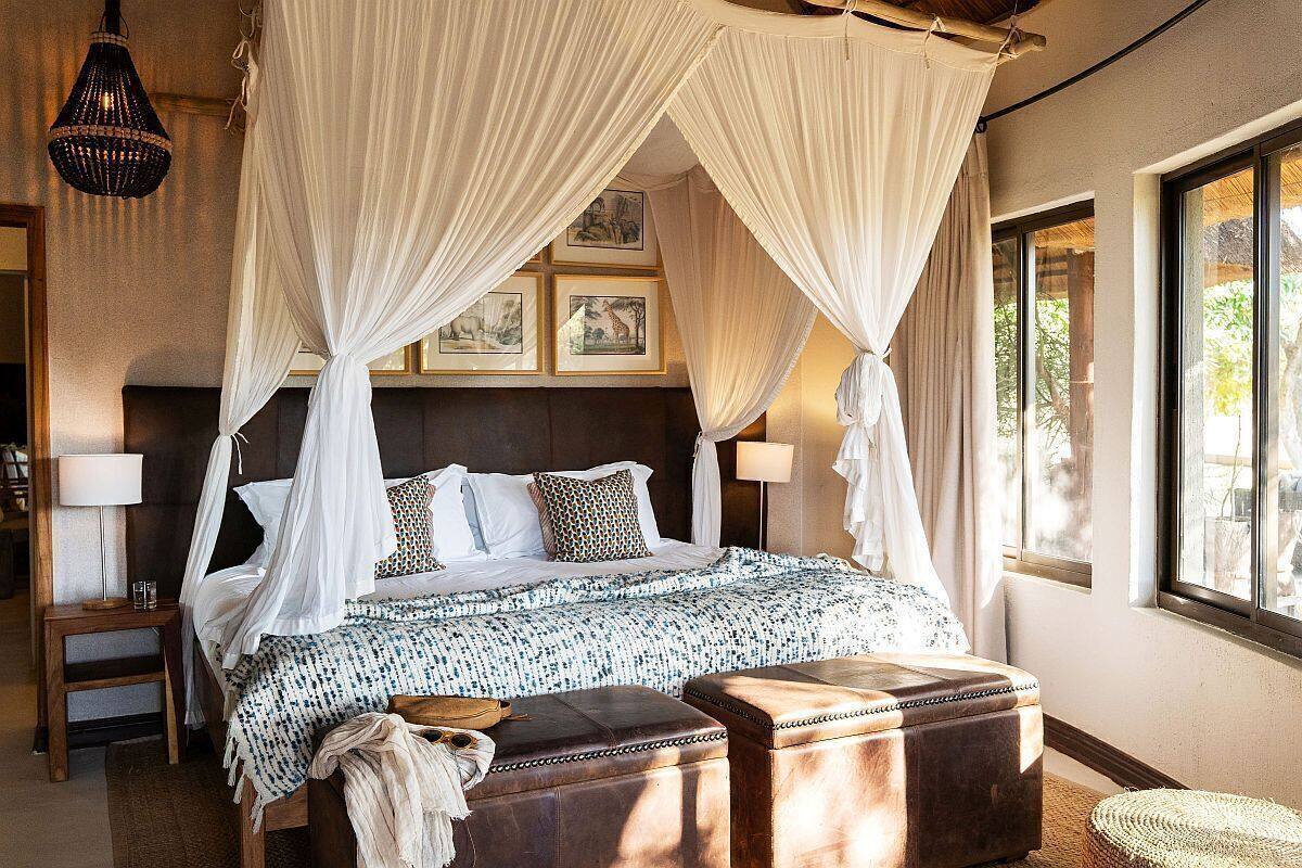 Une chambre à coucher avec un lit à baldaquin, une tête de lit en bois, des lampes de chevet et une vue sur l’extérieur grâce à de grandes fenêtres.