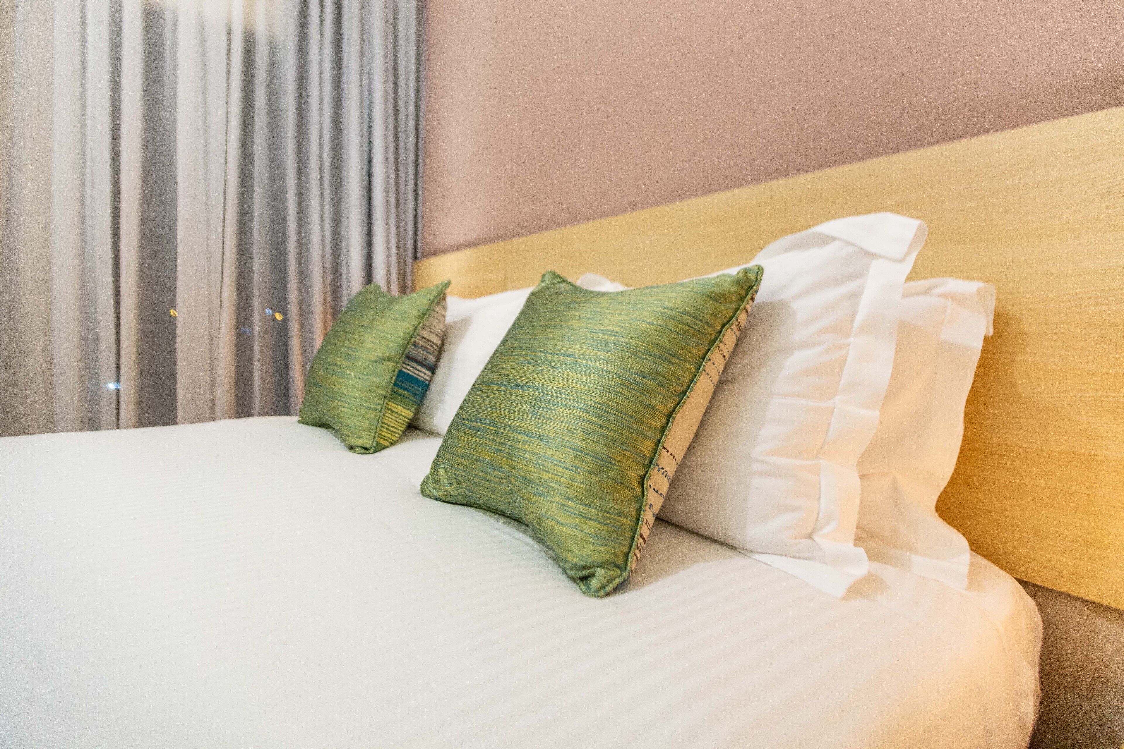 A neatly made bed with white linens and green striped pillows, a wooden headboard, and a window with sheer curtains.