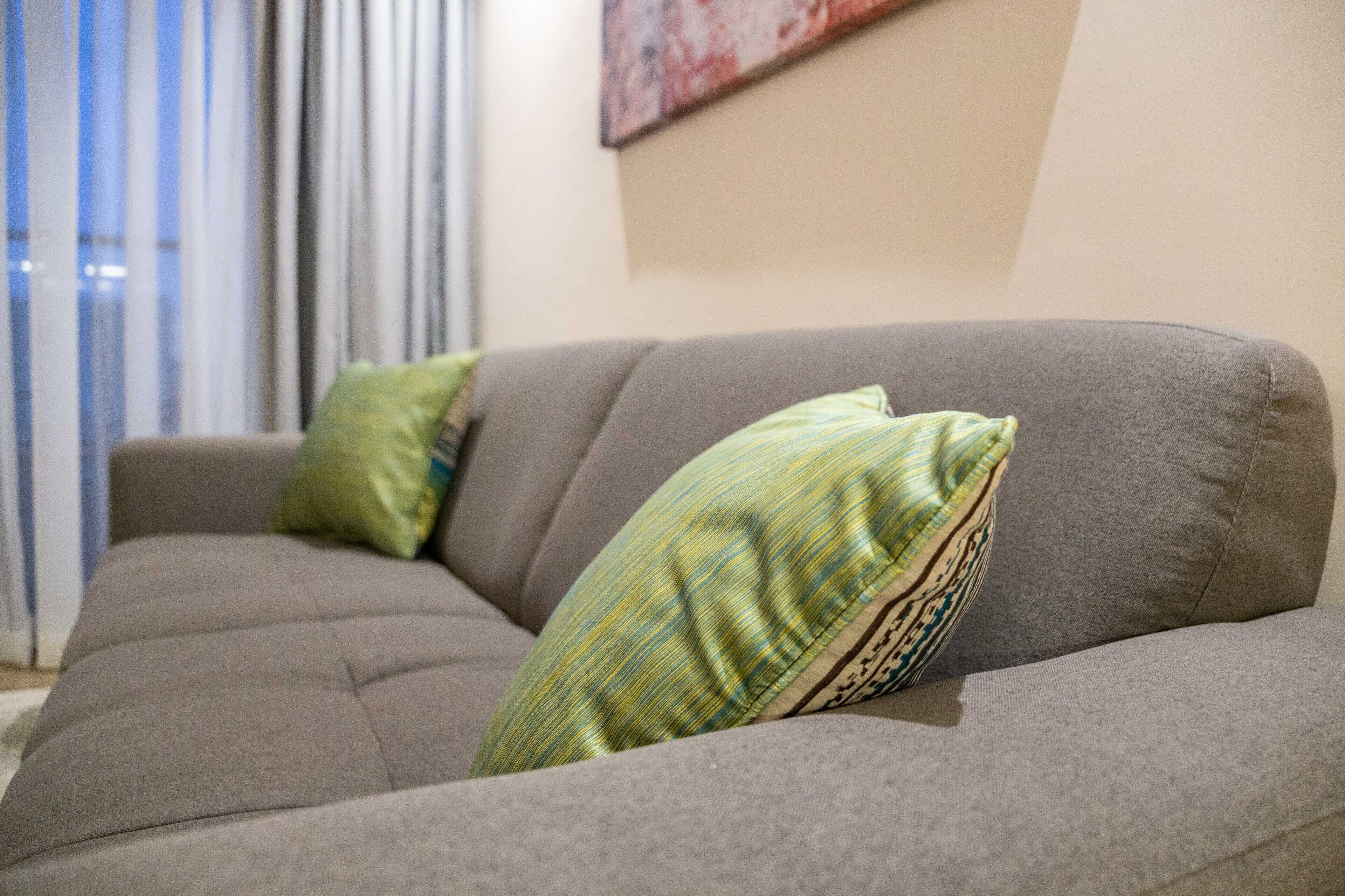 A grey sofa with two green pillows in a room with a window and a painting on the wall.