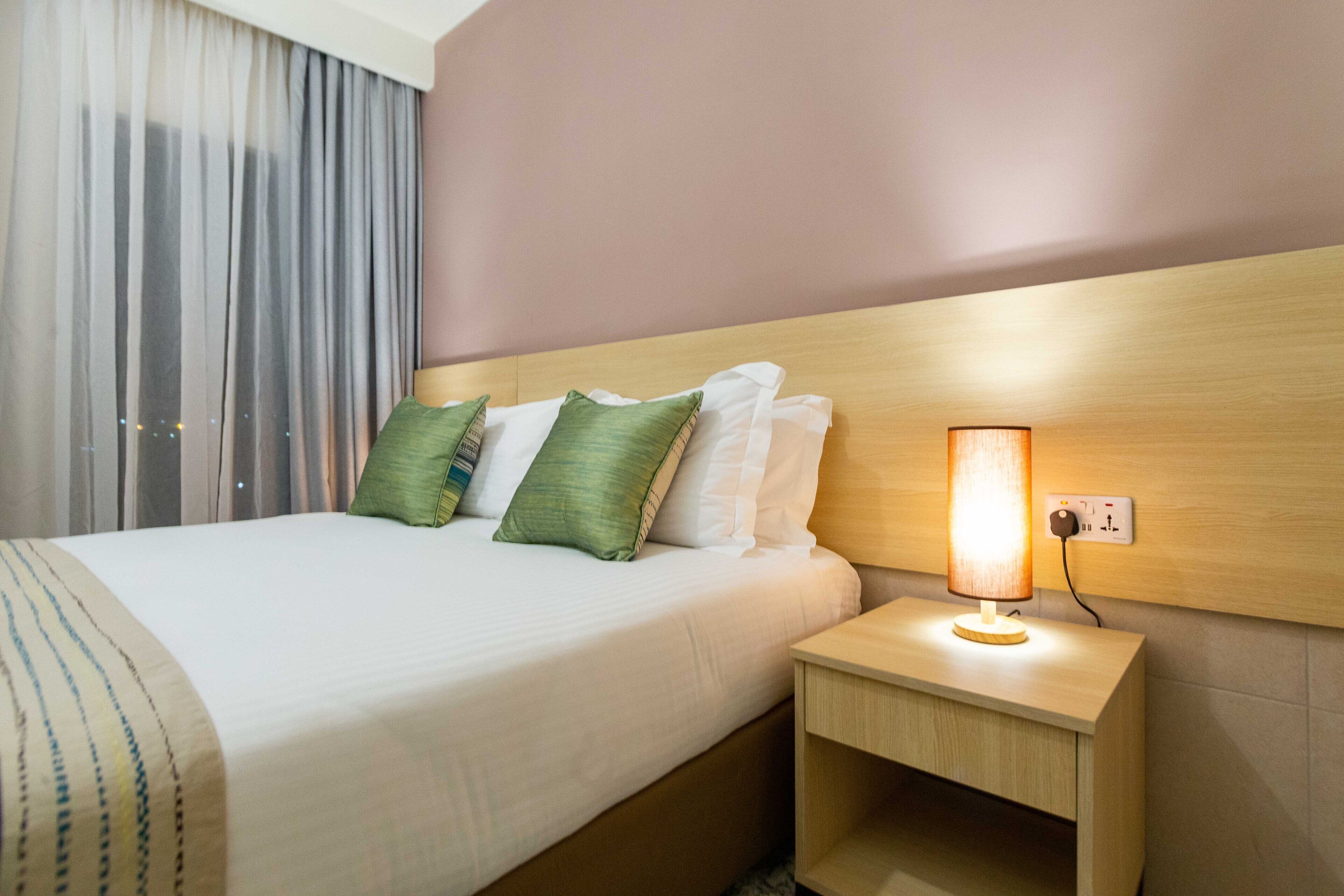 A hotel room with a bed, pillows, a nightstand with a lamp, and a window with curtains.