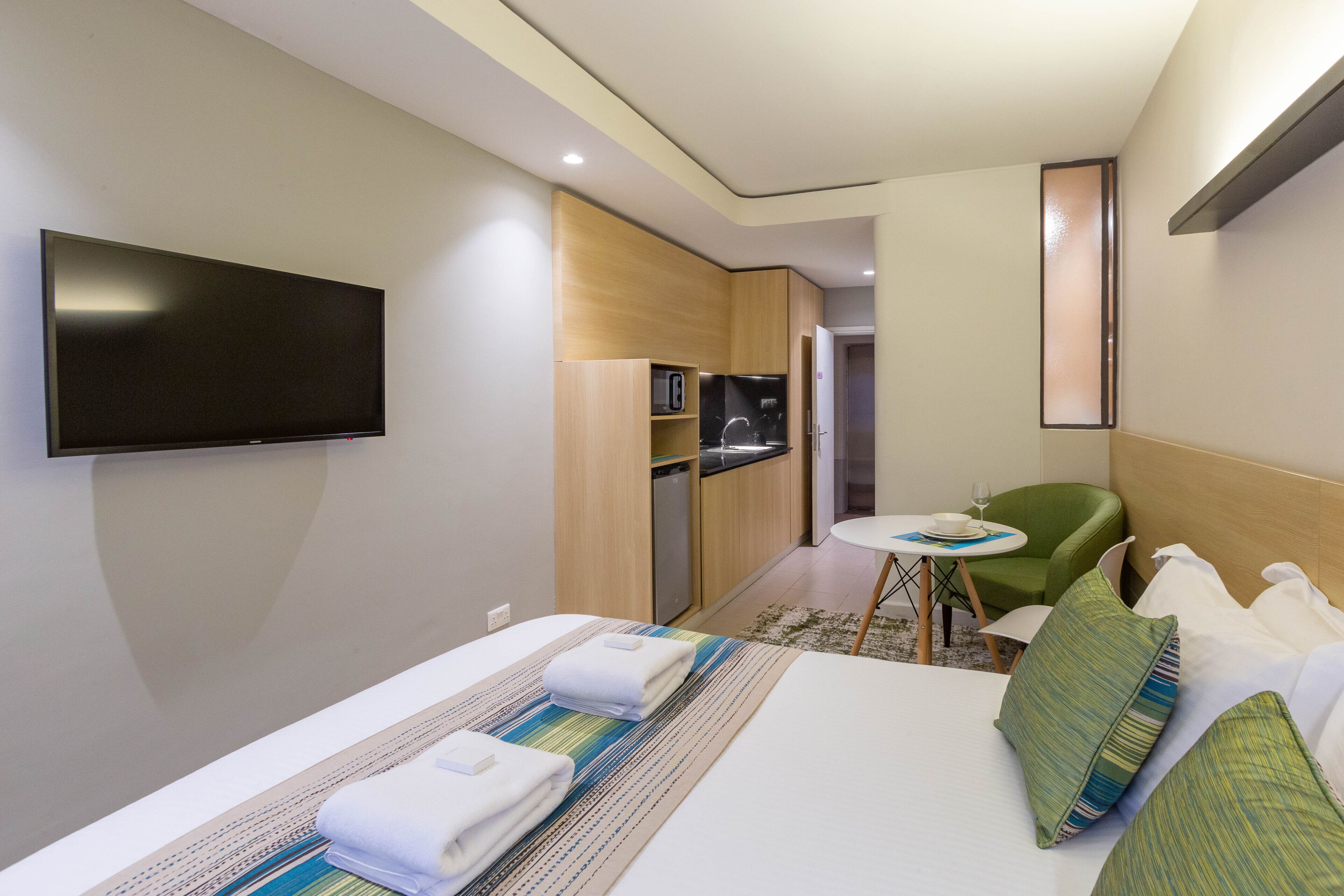 A hotel room with a bed, a TV, a small kitchen area, a dining table, and a green chair.