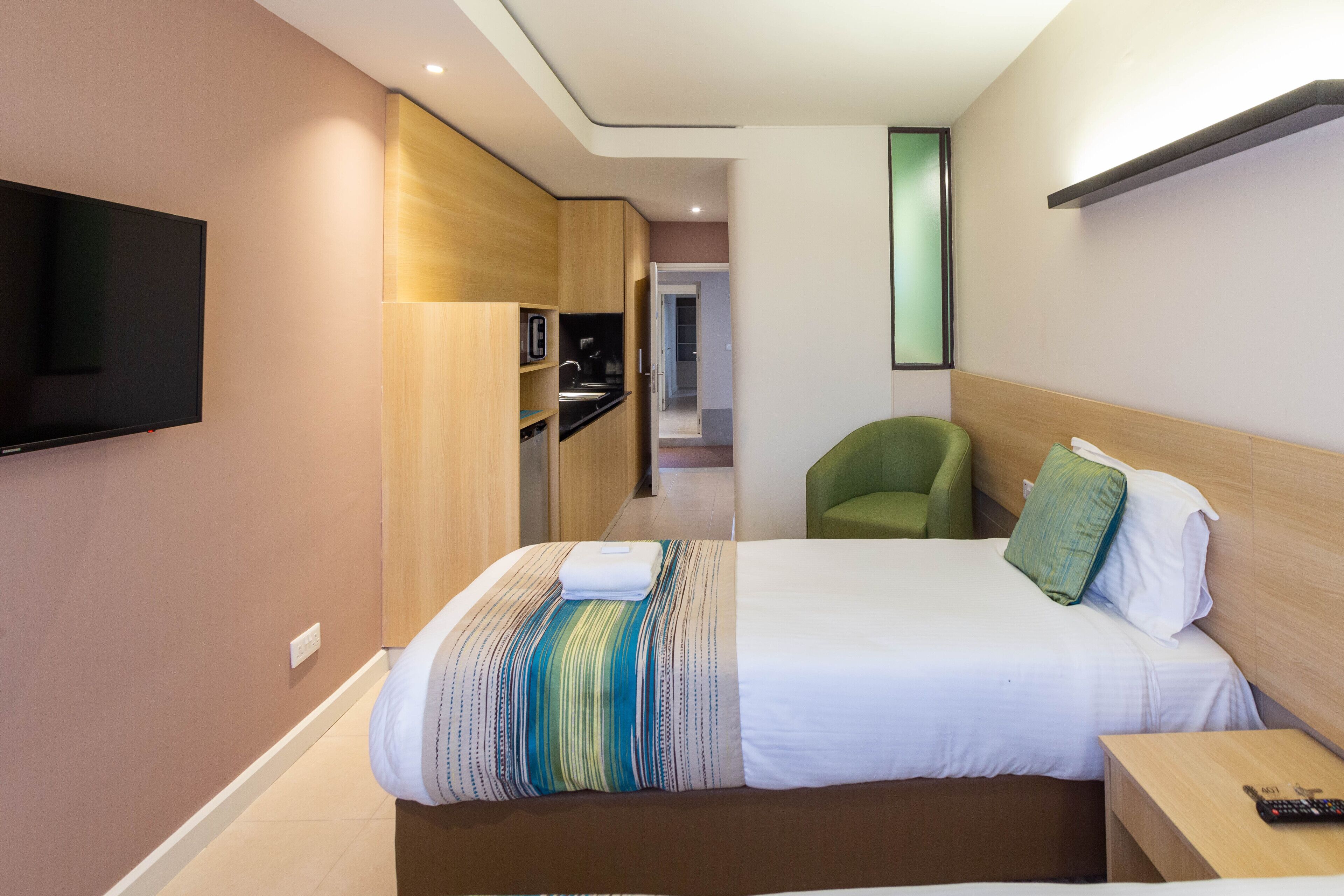 A hotel room with a bed, a TV mounted on the wall, a green chair, and a wooden cabinet.