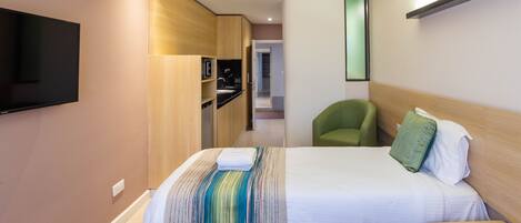 A hotel room with a bed, a TV mounted on the wall, a green chair, and a wooden cabinet.