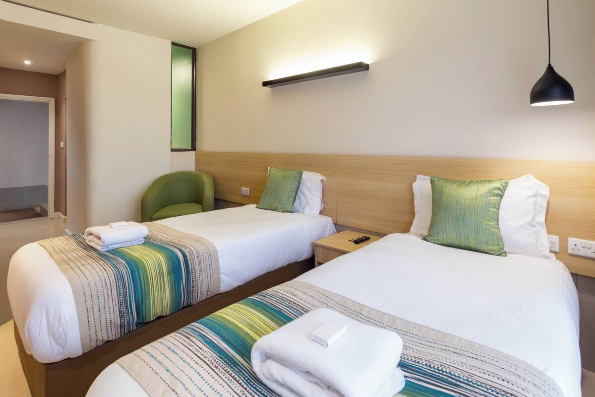 A hotel room with two beds, a green chair, a nightstand, and a wall-mounted light fixture.