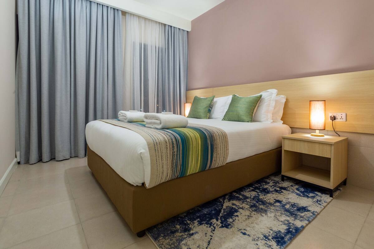 A neatly made bed with a striped blanket, a bedside table with a lamp, and a window with curtains.