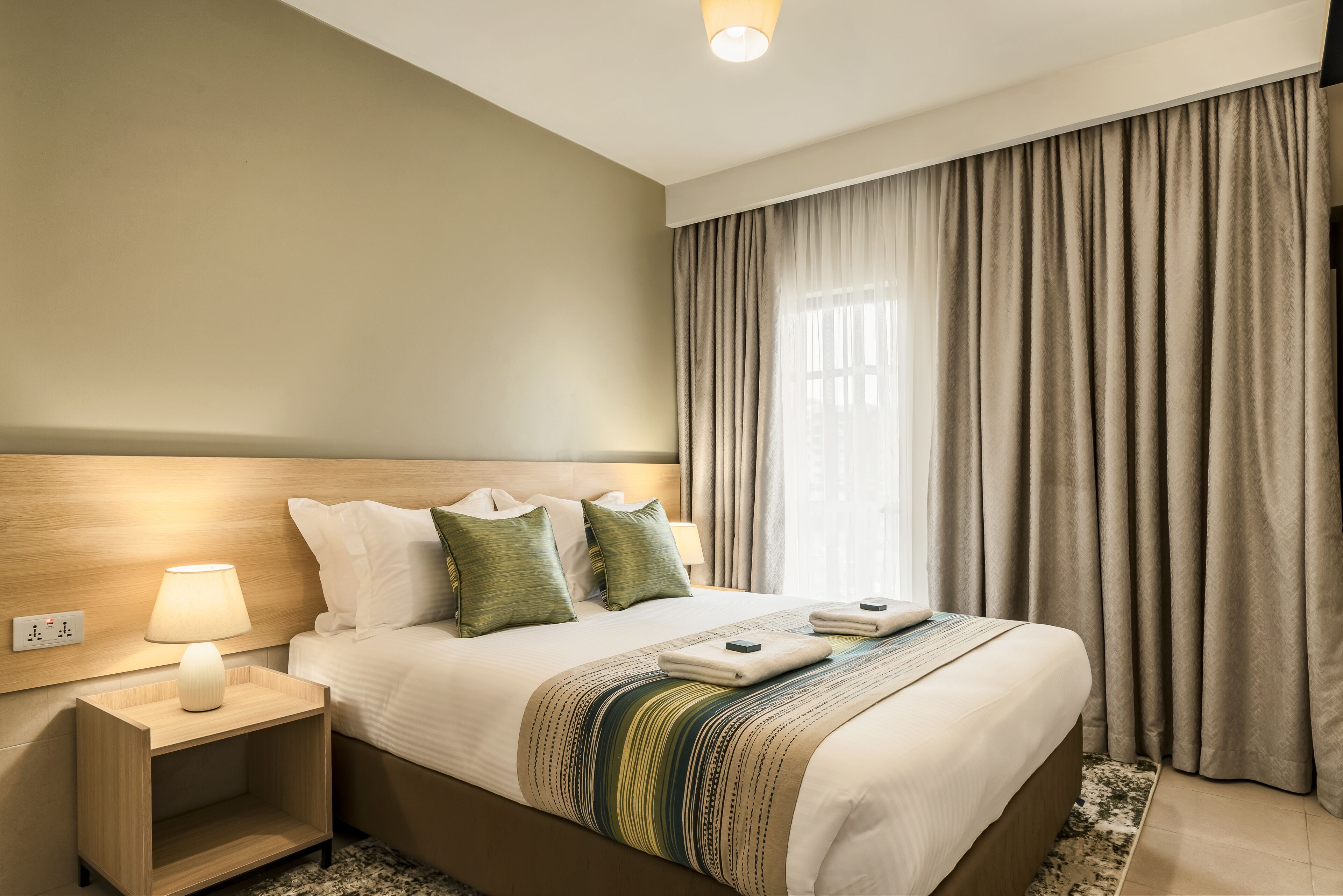 Una habitación de hotel con cama, lámpara de noche, mesita de noche y ventana con cortinas.