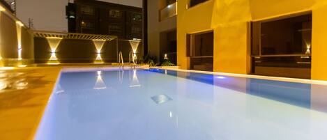 A swimming pool with a clear blue surface, surrounded by a building with multiple windows and balconies.
