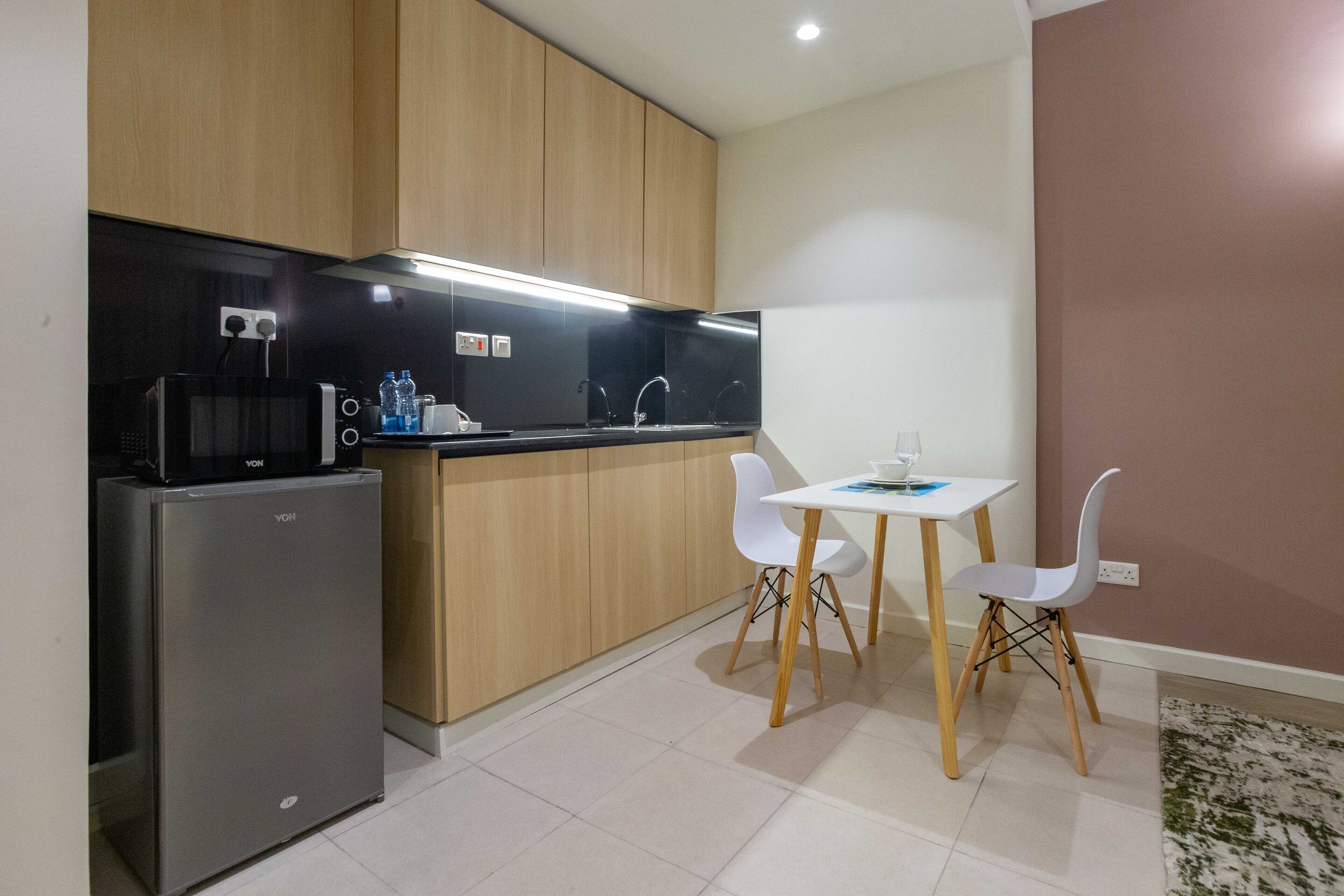 Cocina compacta con refrigerador, microondas y gabinetes de madera. Hay una pequeña mesa de comedor con dos sillas blancas.